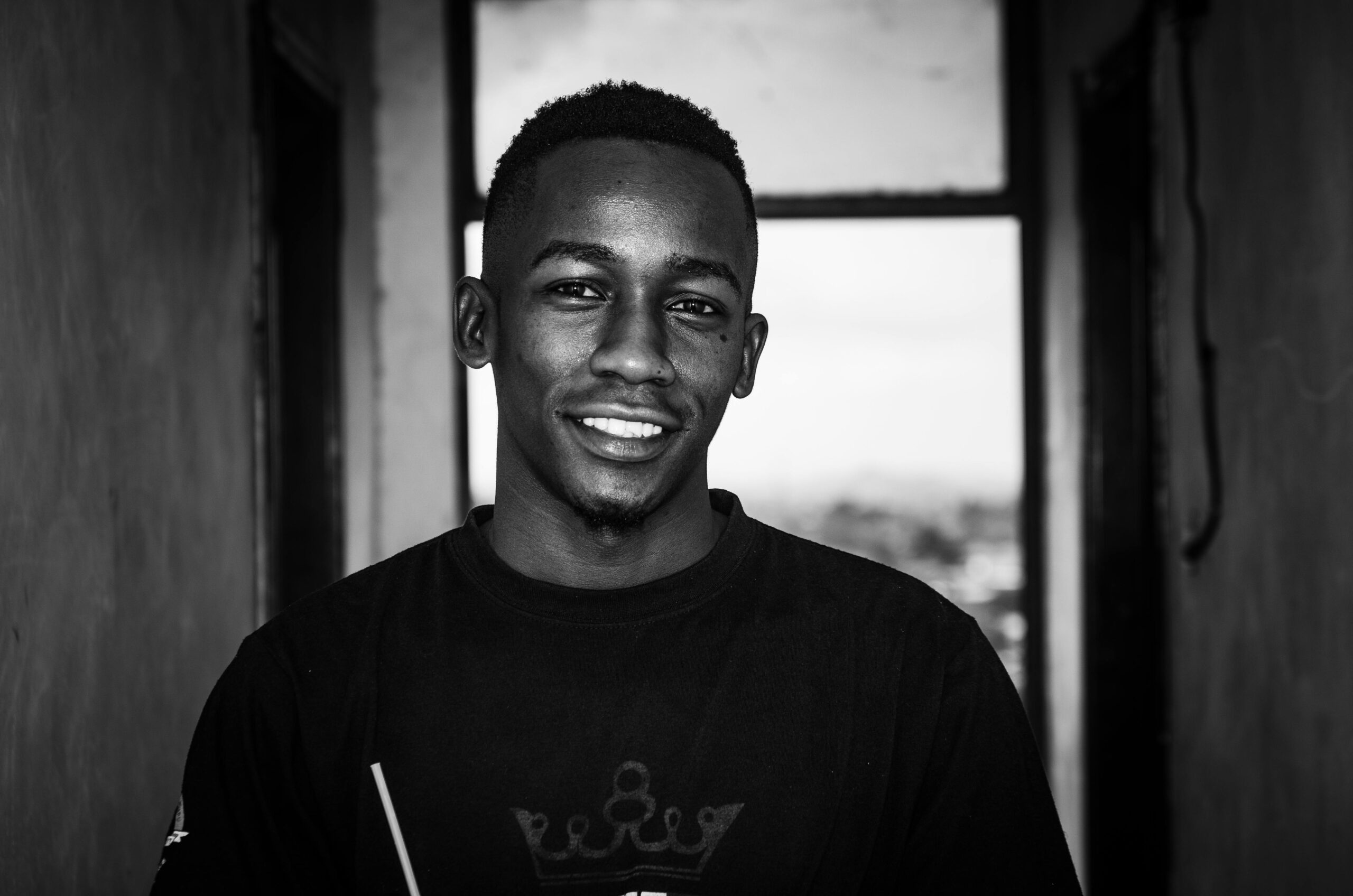 Black and white portrait of a young man smiling, standing in a hallway.